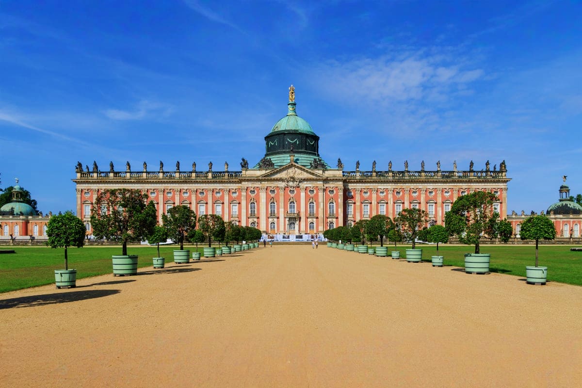Berlin to Potsdam and Sanssouci Palace Day Trip Neues Palais in Potsdam with ornate pink rococo facade, copper dome, manicured gardens, and sand courtyard under blue sky