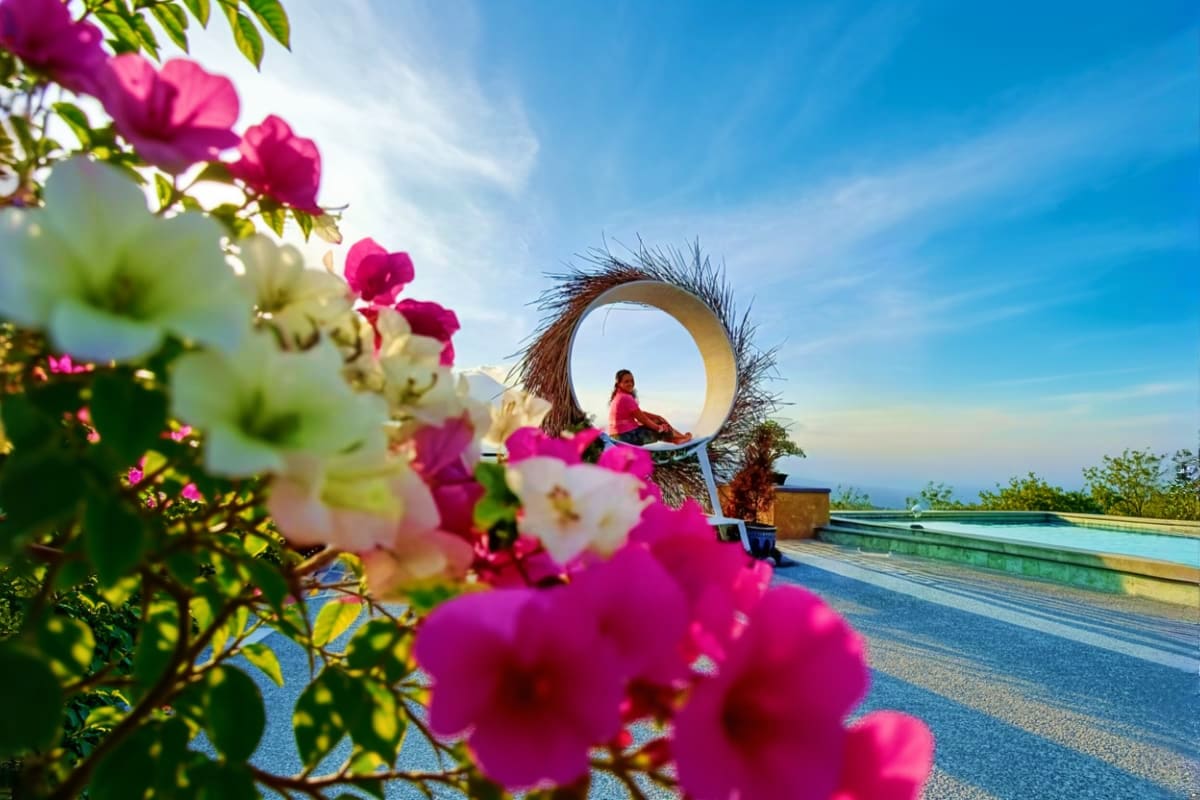 Magenta bougainvillea flowers in foreground with woman sitting in large circular gate structure overlooking Bali landscape an