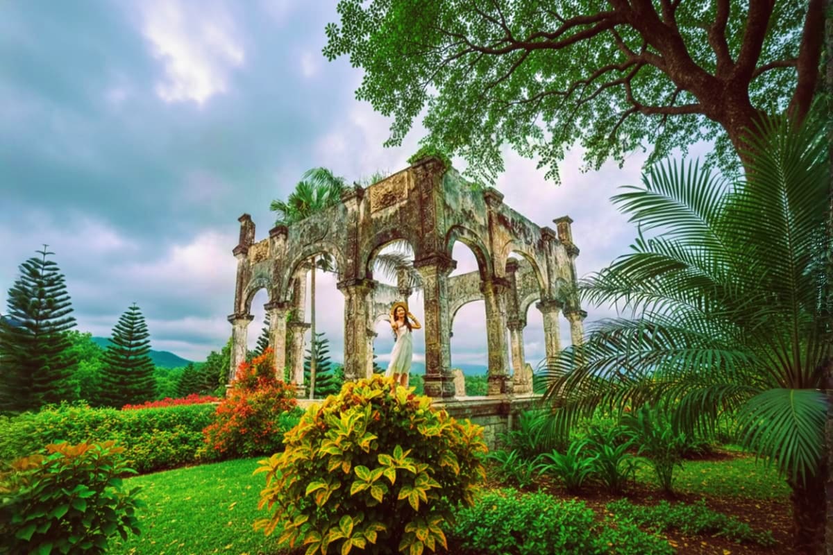 Woman standing in tropical garden with ancient stone archway ruins, palm trees, and mountains in background