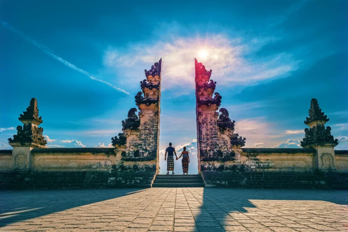 Lempuyang Gate of Heaven Tour Through Bali Most Famous Destinations Couple holding hands standing at the ornate stone gates of Lempuyang Temple with carved towers and red flags against blue sky