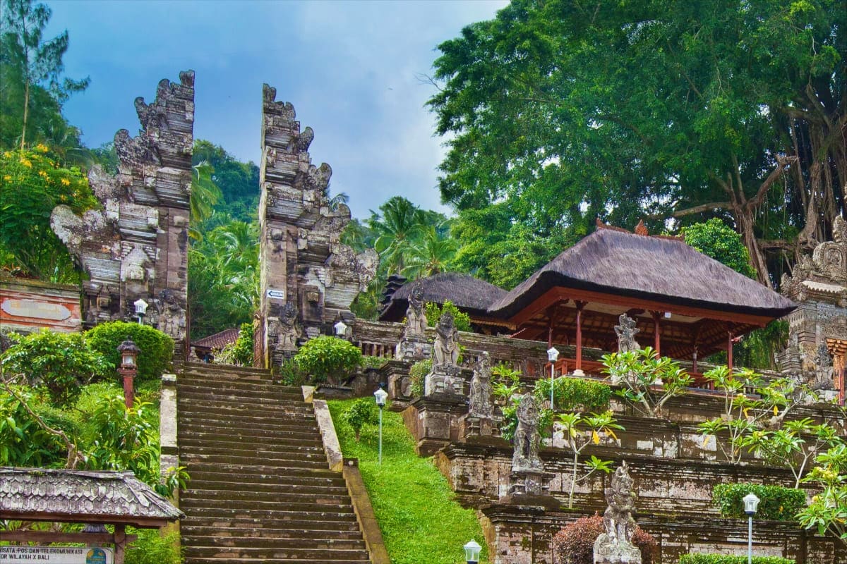 Ancient Balinese temple with ornate stone gates, traditional thatched roof structure, carved statues, and lush tropical veget