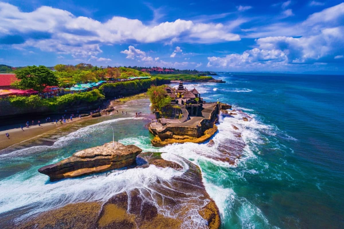 Bali Full Day Private Tour Aerial view of Tanah Lot temple perched on rocky outcrop with crashing waves, beach, and lush green cliffs in Bali