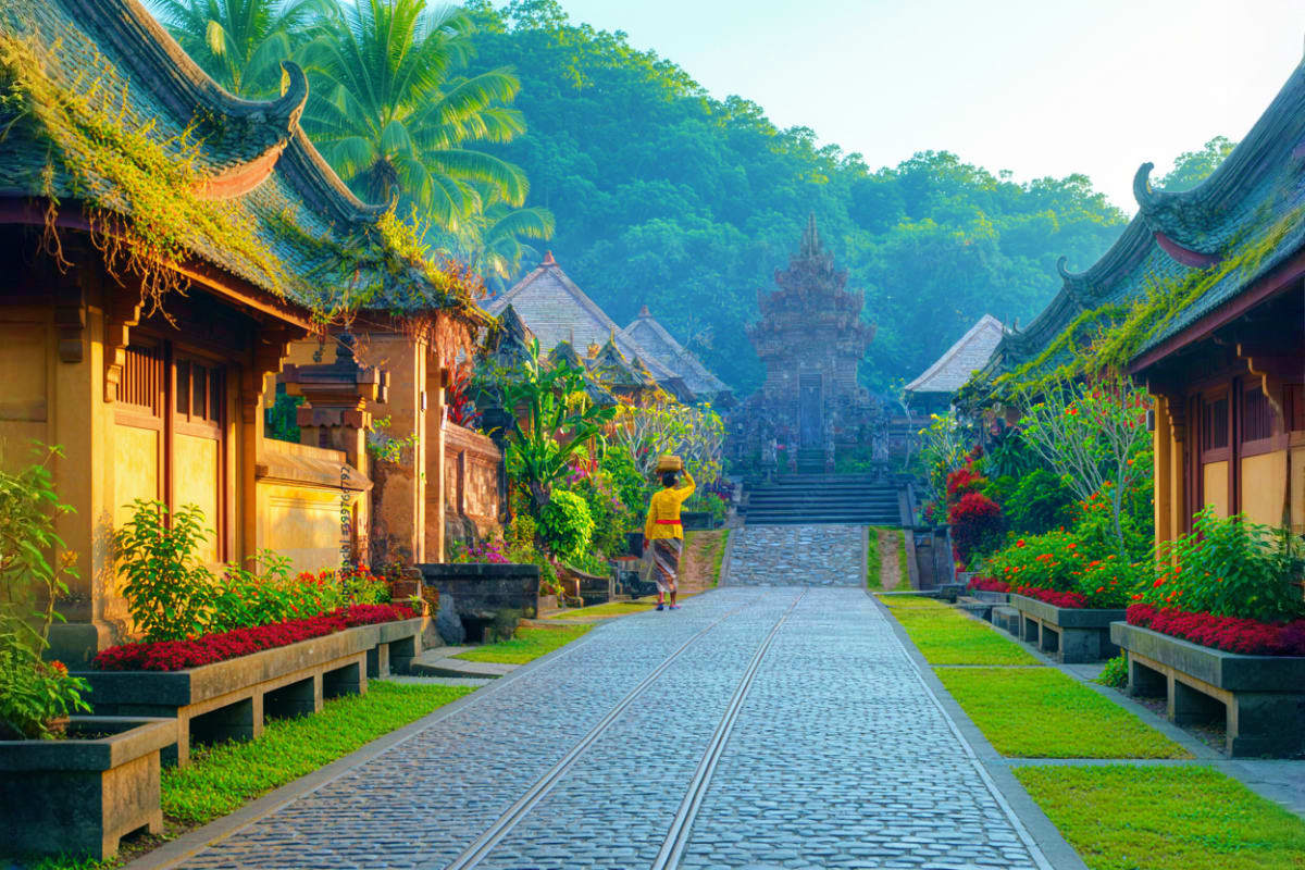Traditional Balinese temple complex with upturned roof eaves, stone pathway, lush tropical gardens, and palm trees creating a