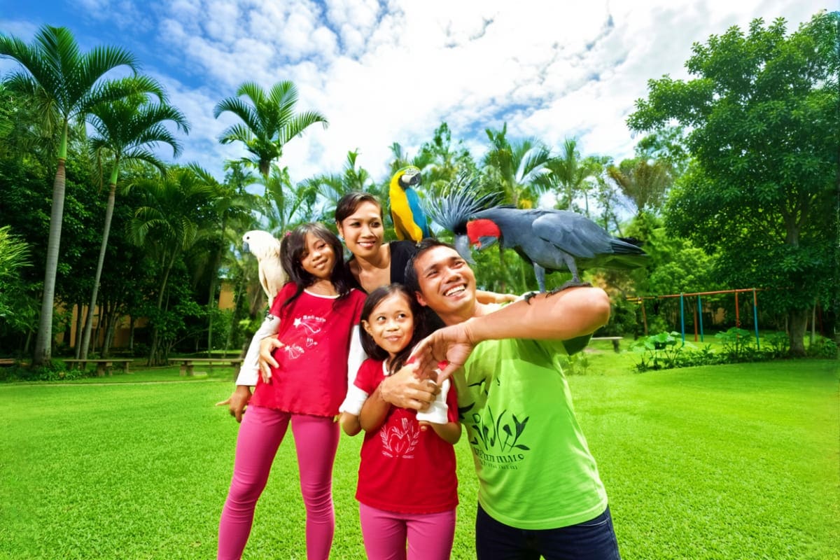 Family holding parrots at Bali Bird Park with tropical birds and palm trees in lush garden setting