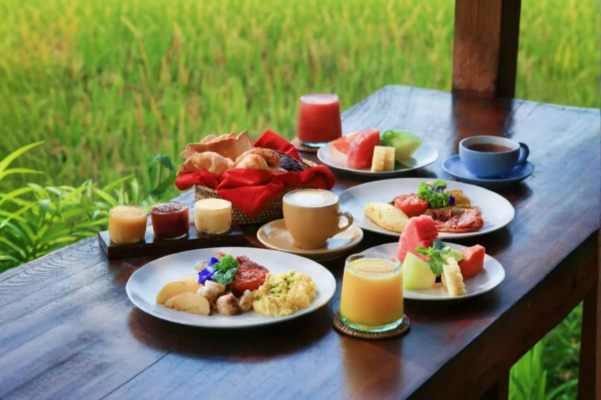 Outdoor breakfast spread at wooden table overlooking rice paddies with eggs, fresh fruit, coffee, juice, and tropical flowers
