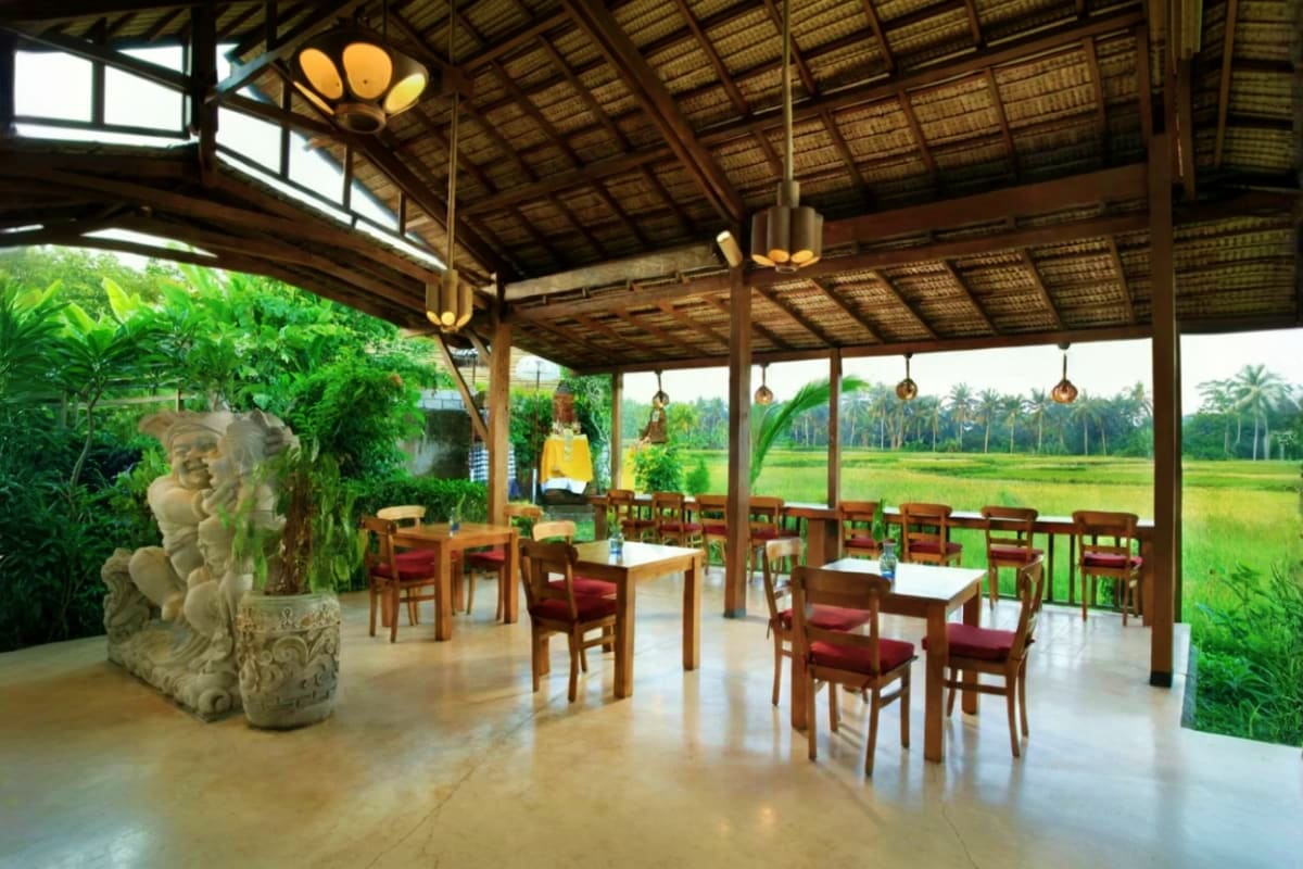 Dining Experience at Bee Cafe Ubud Bali Open-air dining pavilion at Bee Cafe with wooden tables, red chairs, and stone statue overlooking rice paddies in Ubud, Bali