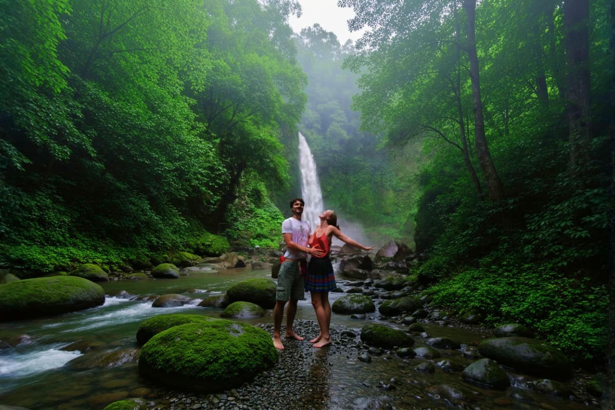 Bali Private Full Day Temple And Waterfall Tour Couple standing on moss-covered rocks viewing a waterfall in a lush green canyon with streaming water and tropical forest in