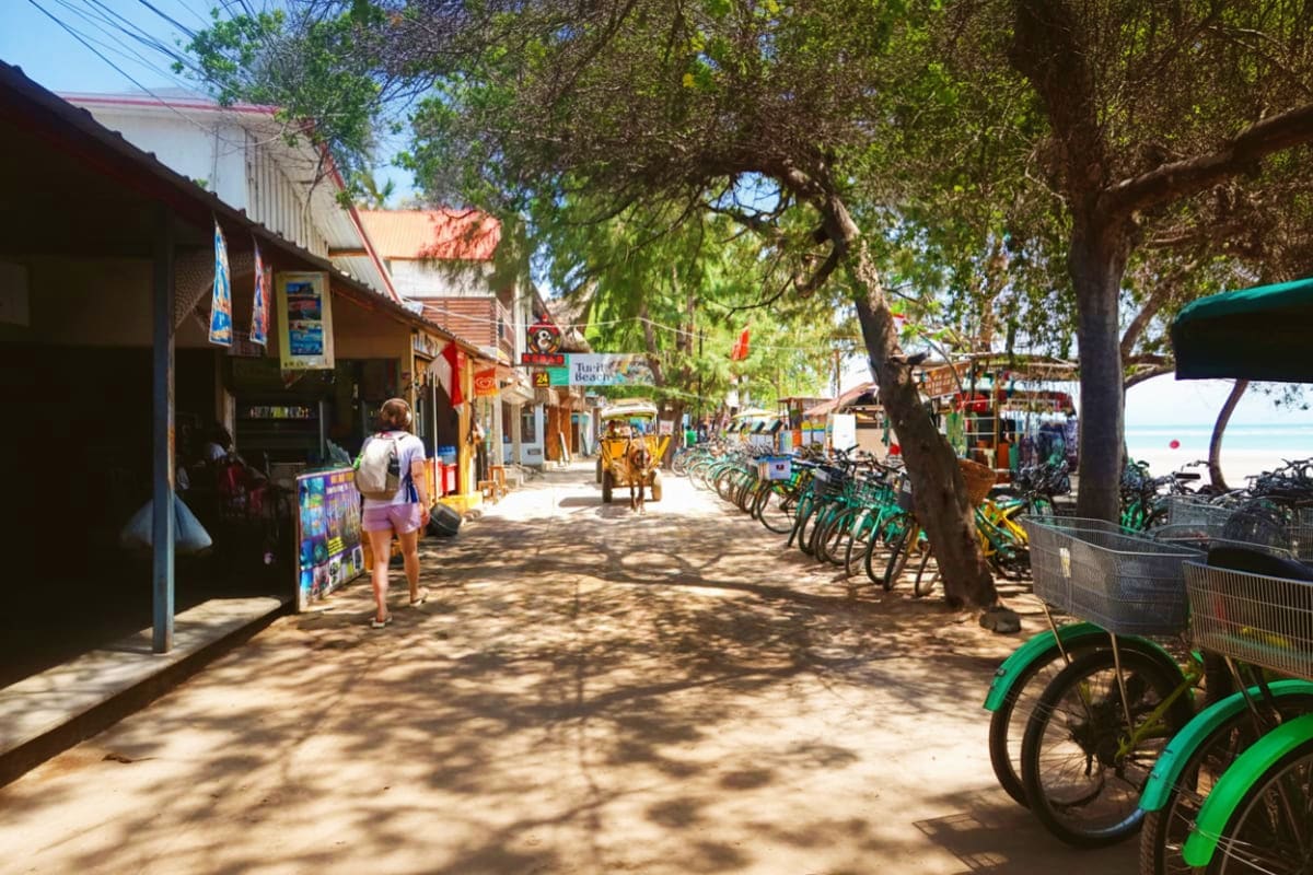 Gili Trawangan and Gili Meno with Gili Air Private Snorkeling Day Tour from Lombok Tropical beach village street with colorful bikes parked along sandy path, shaded by trees, with shops and ocean view in dist