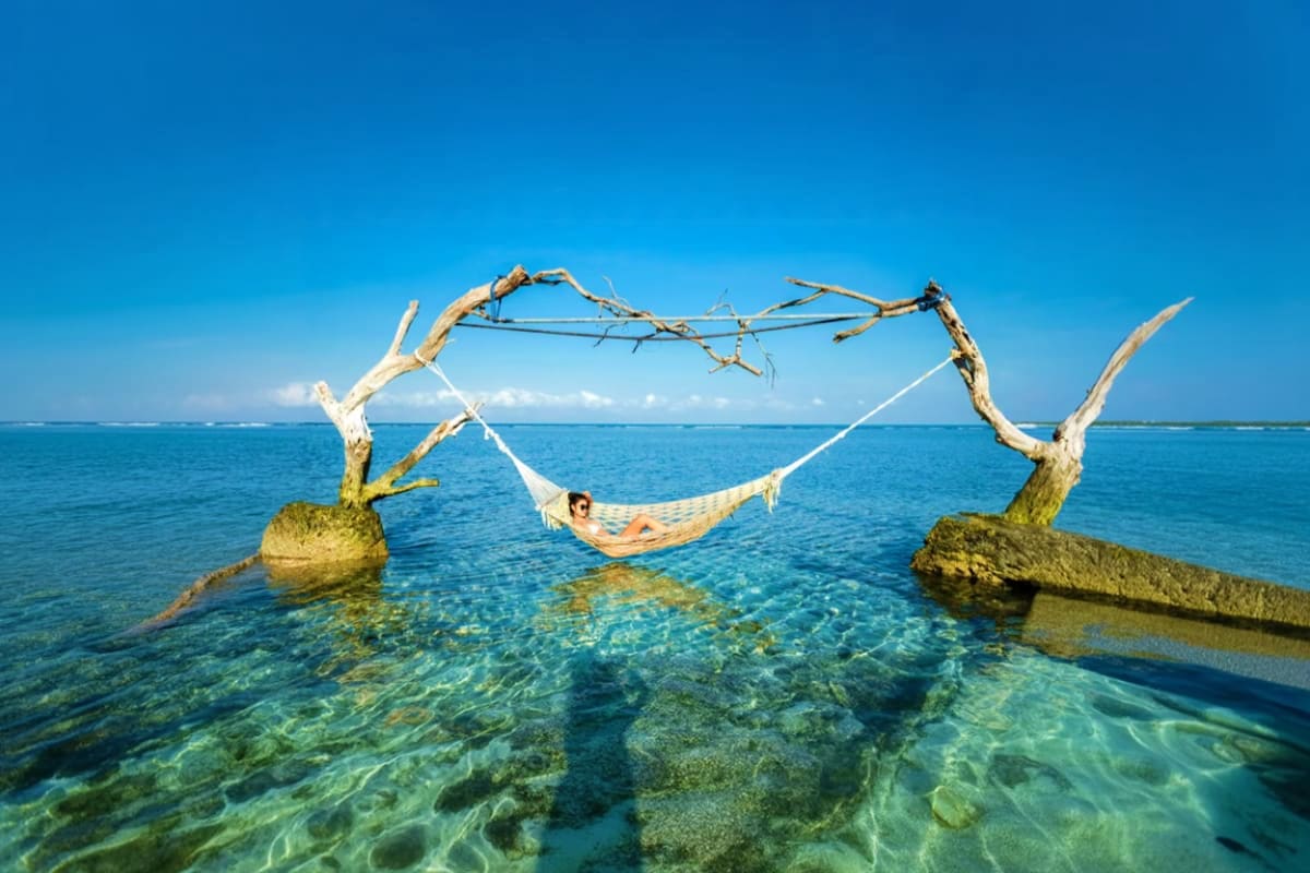 Person relaxing in hammock suspended between driftwood branches in shallow turquoise ocean water off Gili Islands
