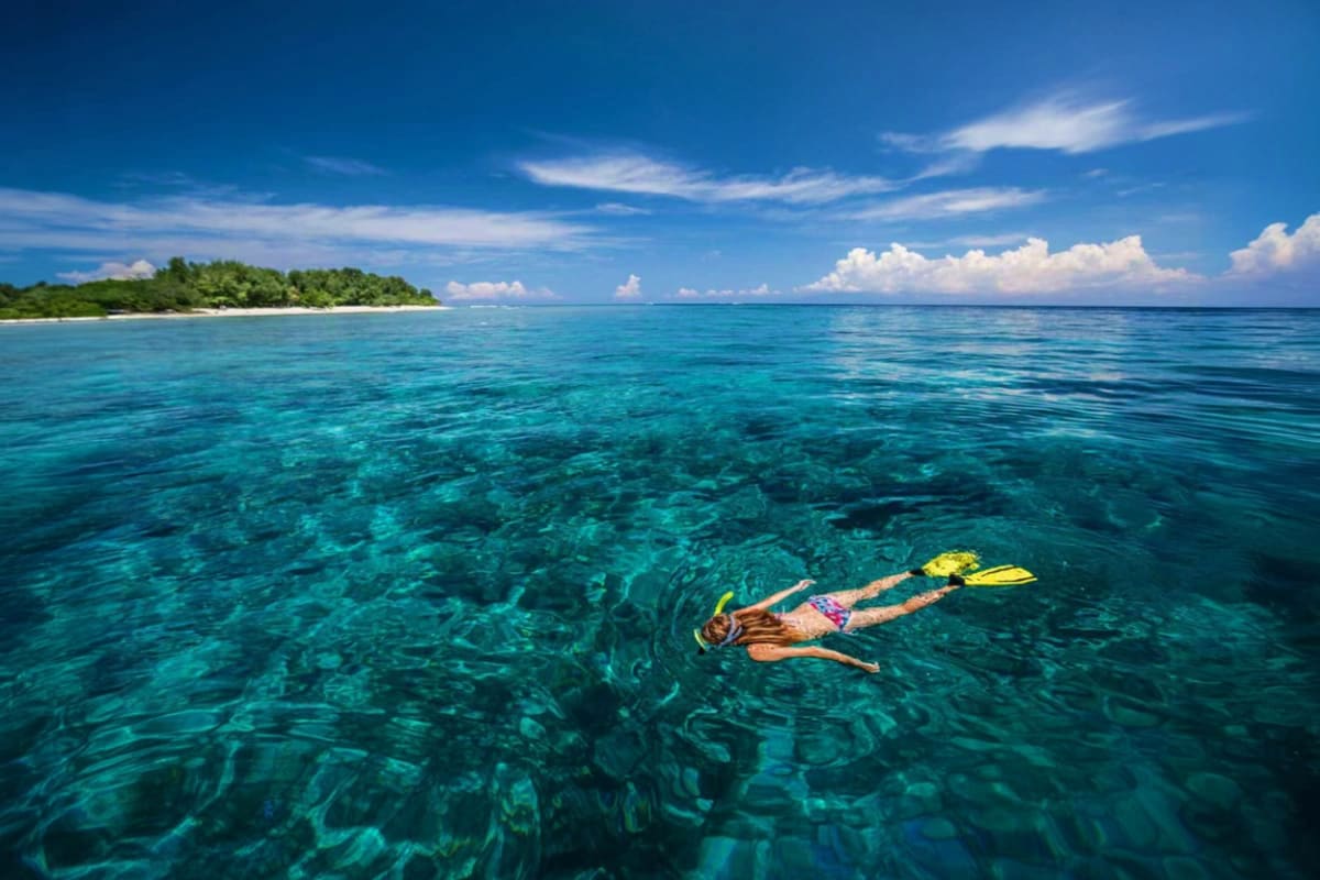 Man snorkeling with yellow fins in crystal clear turquoise water near tropical island with white sand beach and green vegetat