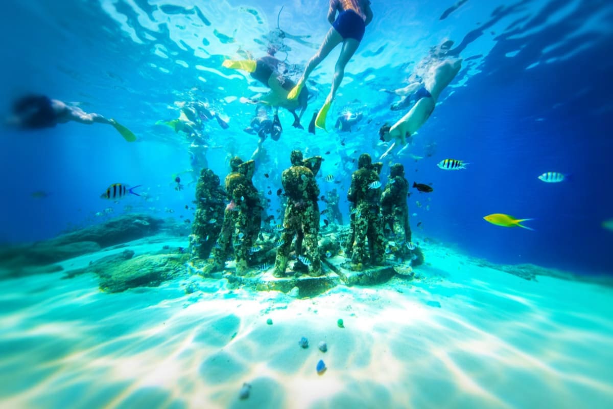 Multiple snorkelers swimming above underwater statue ruins surrounded by tropical fish and coral on sandy ocean floor