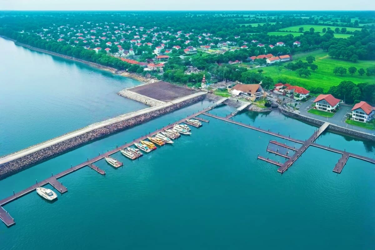 Aerial view of Sanur Harbor with moored boats, wooden docks, breakwater, and coastal village surrounded by green fields and t