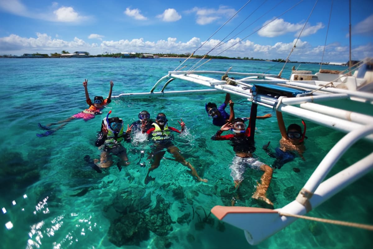 Mactan Water Sports Activities in Cebu Group of snorkelers in turquoise water next to white outrigger boats with clear sky and tropical shoreline in background
