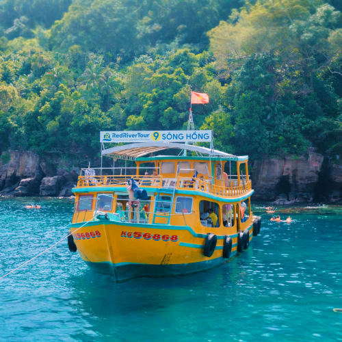 Bright yellow tour boat on turquoise water with lush green forested cliffs and red flag in background