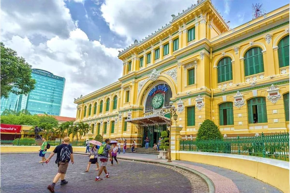 Yellow colonial-era building with clock tower and green shutters, tourists walking in plaza, Ho Chi Minh City landmark