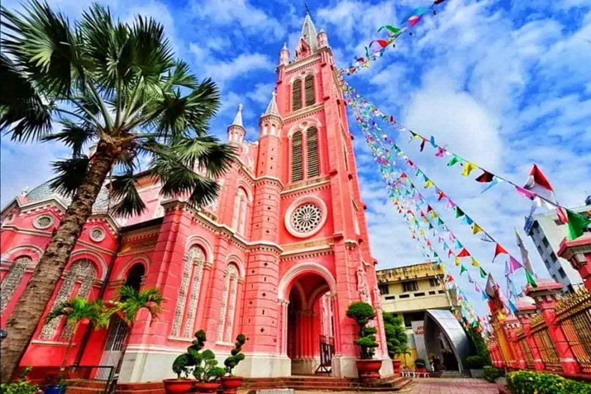 Bright pink Gothic cathedral with tall spire in Ho Chi Minh City, decorated with colorful prayer flags and palm trees