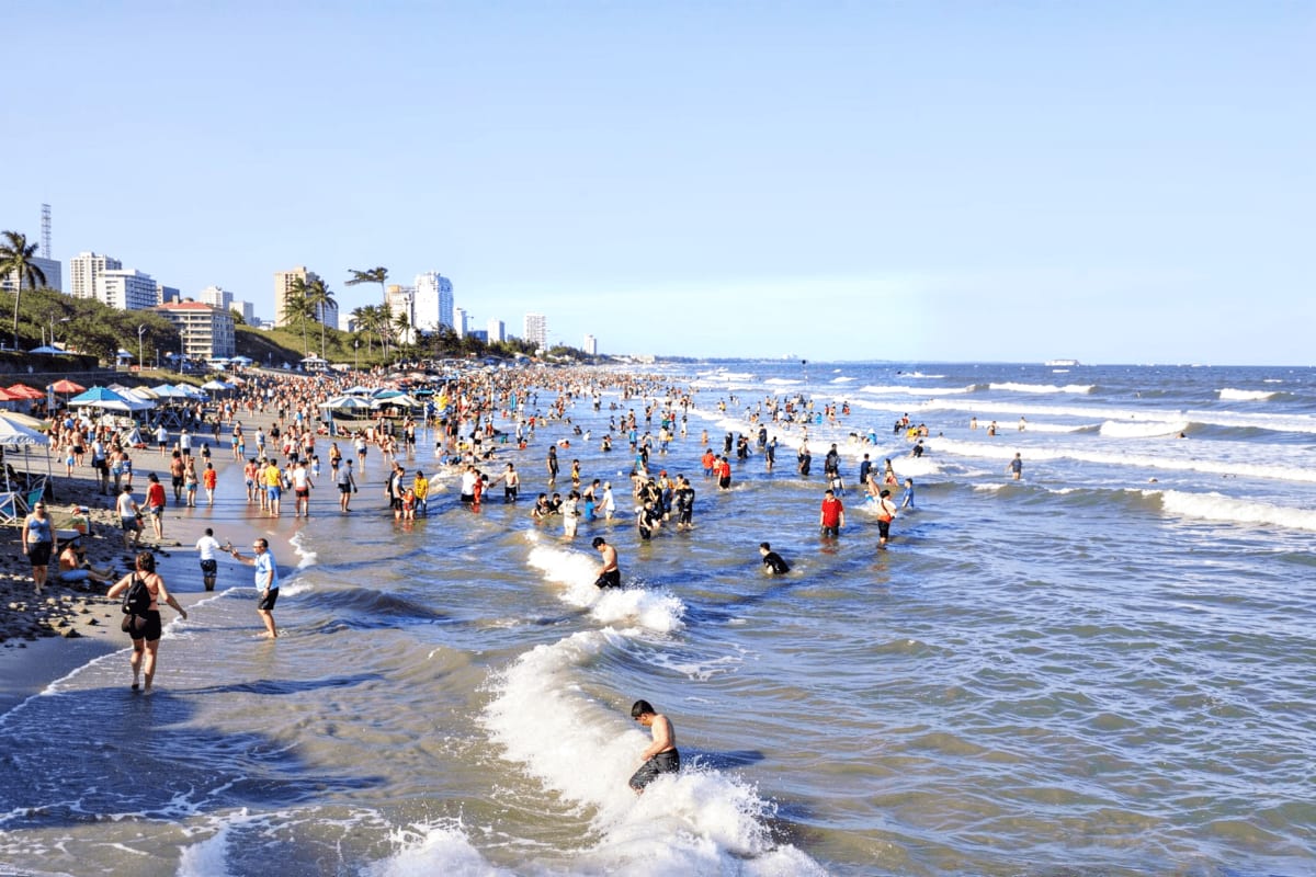 Vung Tau beach Private Tour Full Day Crowded beach scene with swimmers and beachgoers in the ocean waves, sandy shore, palm trees, and high-rise buildings along t