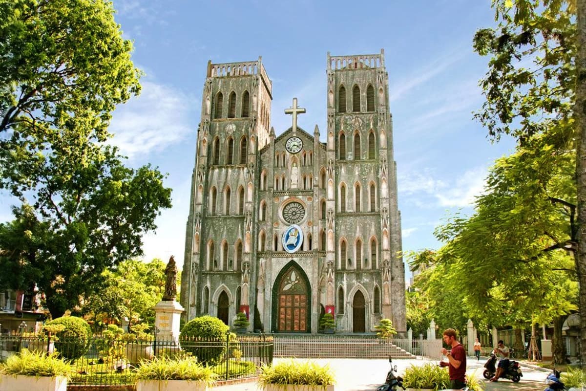 St. Joseph's Cathedral in Hanoi with two tall Gothic towers, cross at top, clock face, surrounded by green trees and visitors