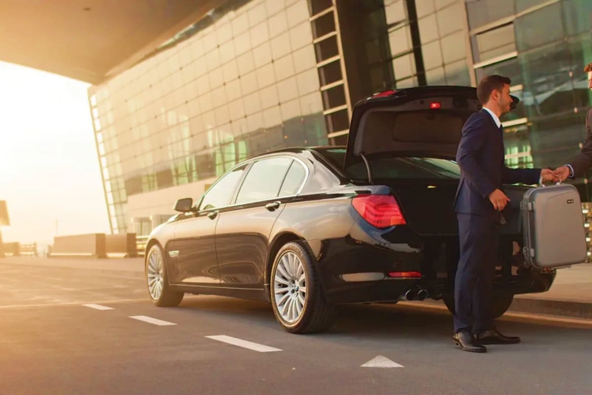 Businessman in suit standing beside black sedan with open trunk, holding luggage at modern office building entrance