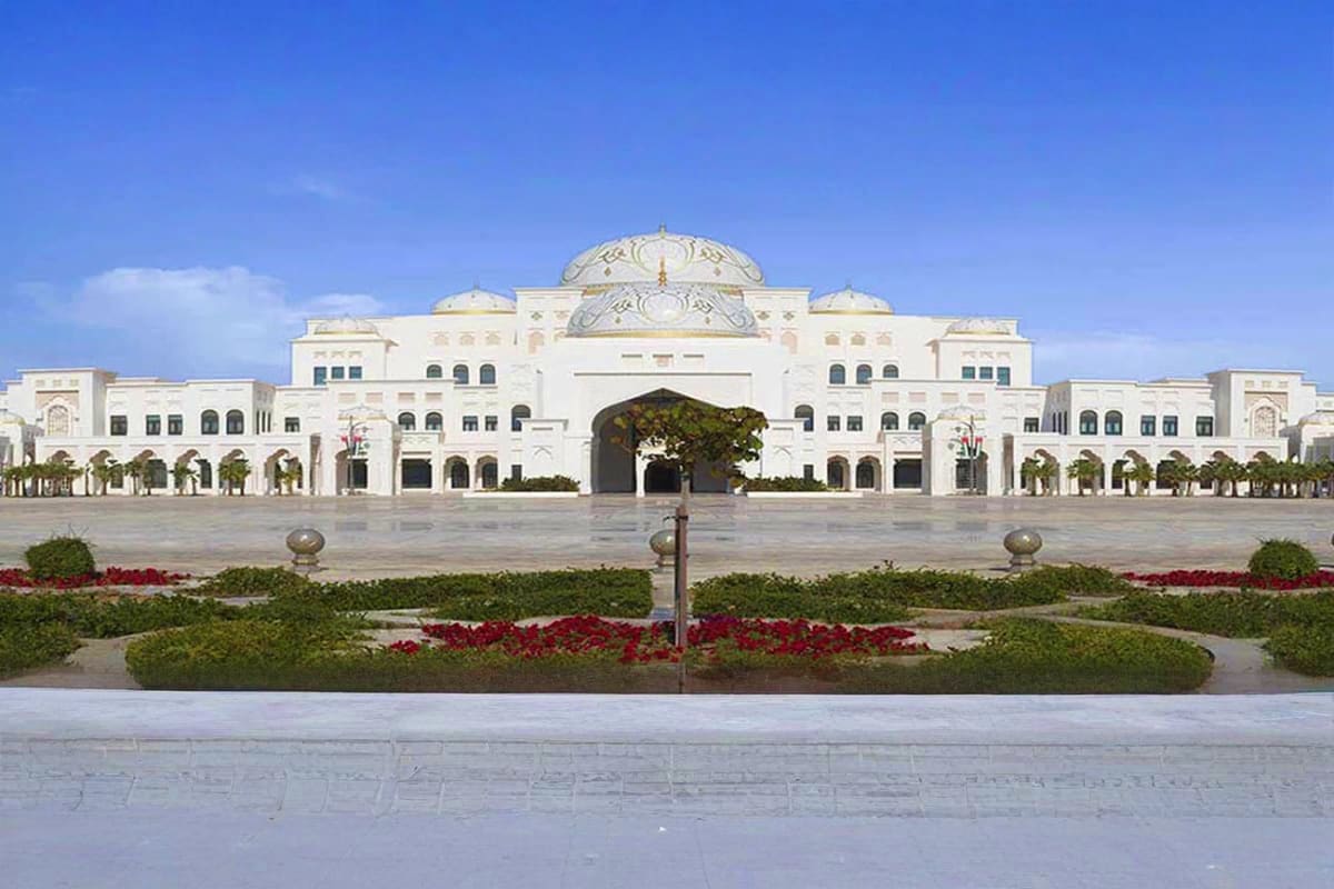 White neoclassical building with domed roof and symmetrical wings, featuring manicured gardens with red flowers in foreground