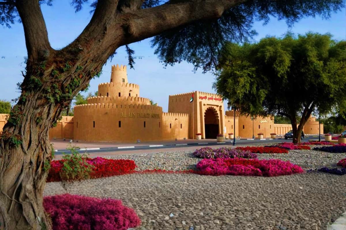 Al Ain Palace Museum with ornate mud-brick facade, crenellated towers, and red flowering plants in foreground under clear sky