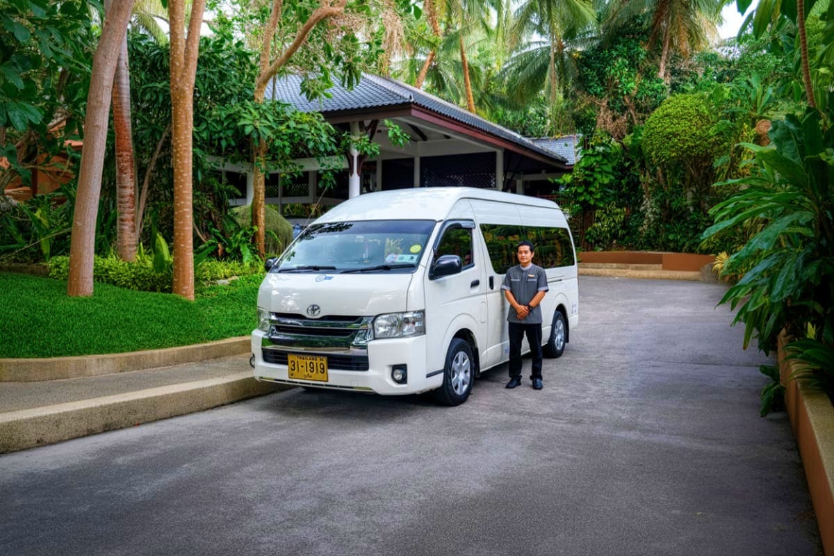 Bangkok Airport to Hua Hin Hotel White Toyota van with driver standing beside it in tropical resort driveway with palm trees and modern building