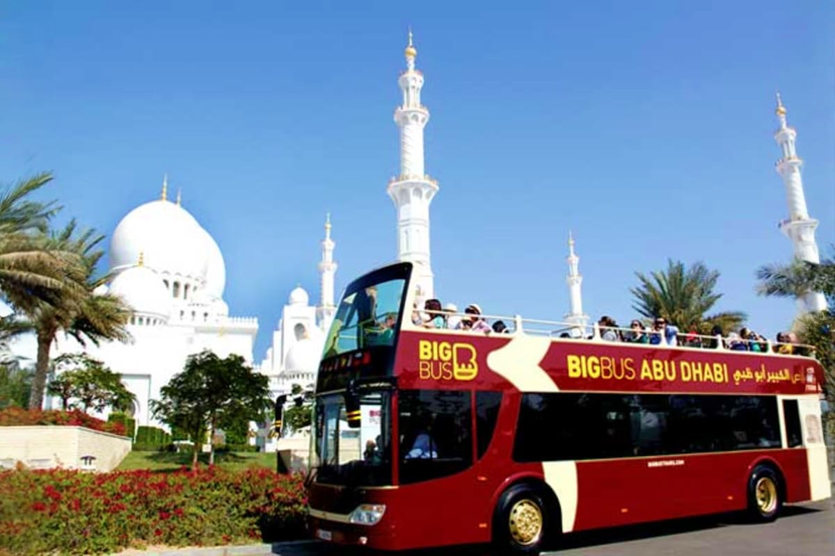 Red and cream Big Bus Abu Dhabi double-decker sightseeing tour bus passing Sheikh Zayed Grand Mosque with white domes and min