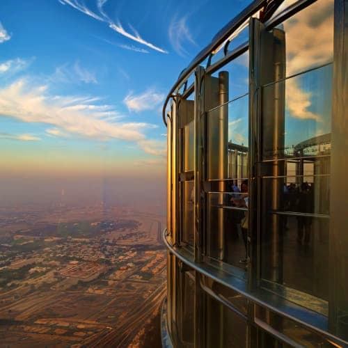 View from Burj Khalifa observation deck showing curved windows, blue sky with clouds, and cityscape landscape far below