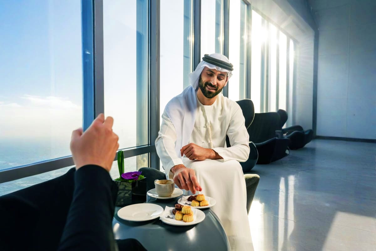 Arab man in traditional white dishdasha dining with pastries in high-rise cafe overlooking Dubai cityscape and ocean view fro