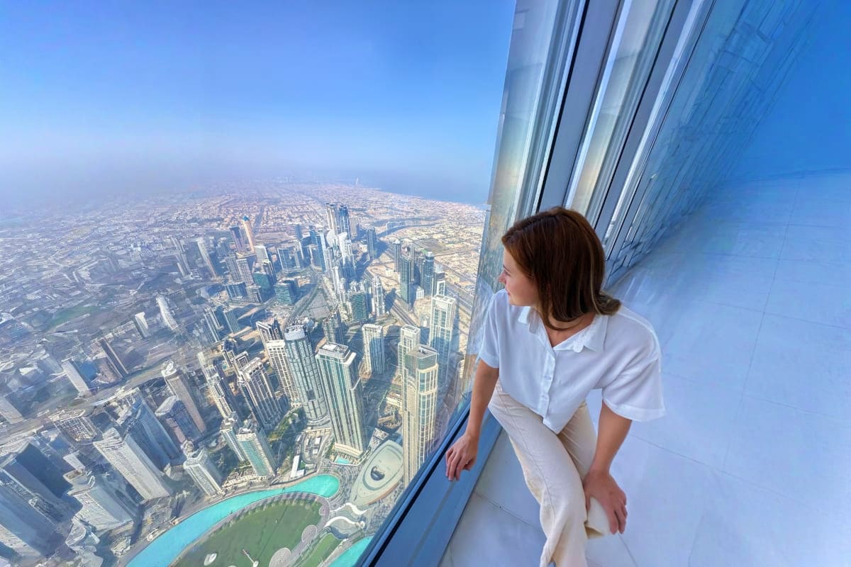 Woman looking out from Burj Khalifa observation deck at Dubai cityscape with turquoise fountain below