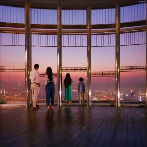 Family viewing cityscape from floor-to-ceiling windows at high-altitude observation deck overlooking Dubai at sunset