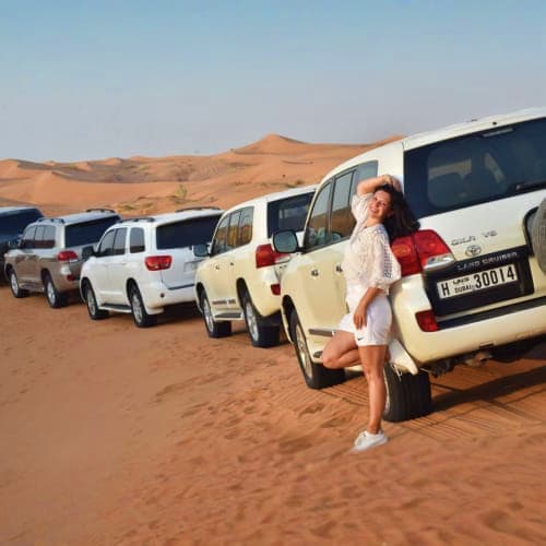 Woman standing beside white SUV parked in desert sand dunes with additional vehicles lined up in background