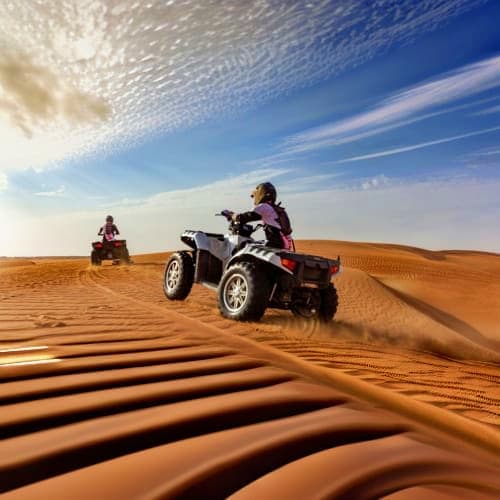 Two riders on quad bikes traversing golden sand dunes under blue sky with wispy clouds during desert safari adventure.