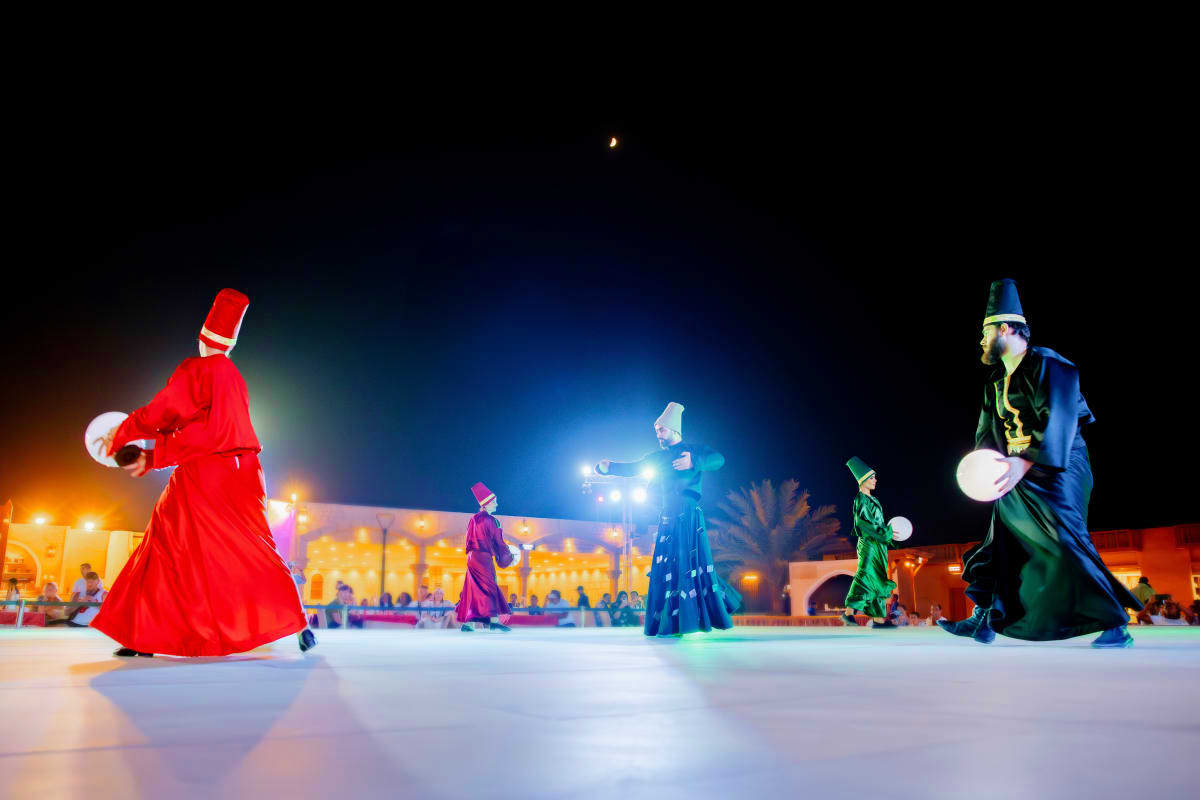 Performers in colorful traditional costumes juggling at night in a Moroccan desert plaza with illuminated buildings and palm