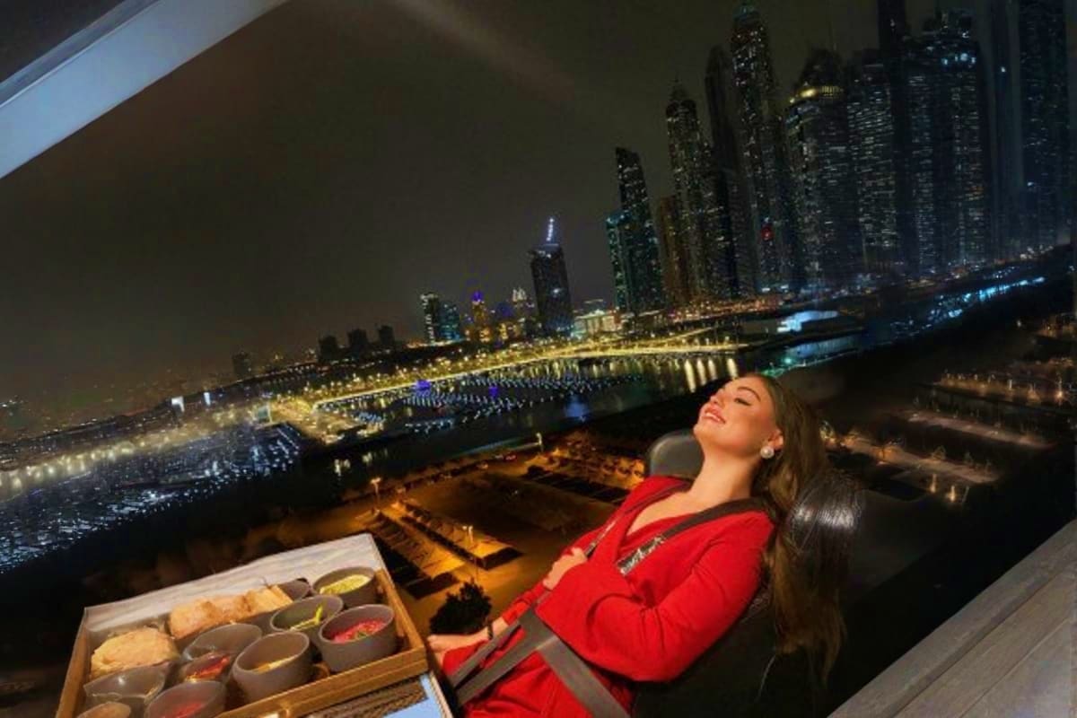 Woman in red jacket enjoying dinner with city skyline and illuminated waterfront buildings at night from elevated dining plat