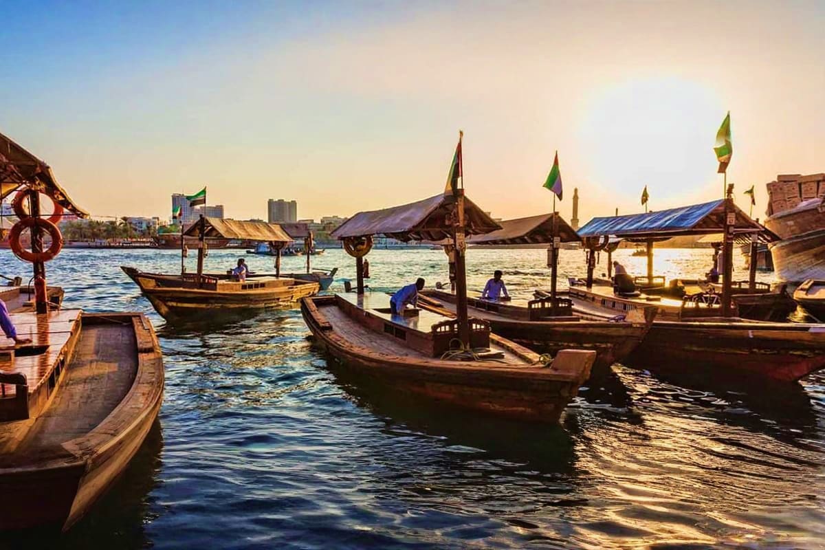 Dubai City Tour From Abu Dhabi Traditional wooden abra boats docked at sunset in Dubai Creek with historic architecture and city skyline in background