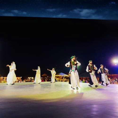 Dubai Desert Safari with Dune Bashing Group of performers in traditional white dress dancing on illuminated stage under night sky at desert event in Dubai
