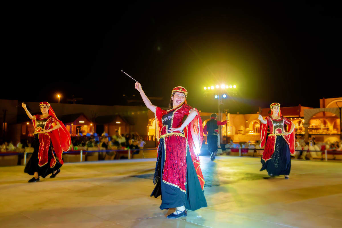 Dubai Desert Safari with Dune Bashing Female performers in traditional red and blue Emirati costumes dancing at night in a desert village square with audience seat