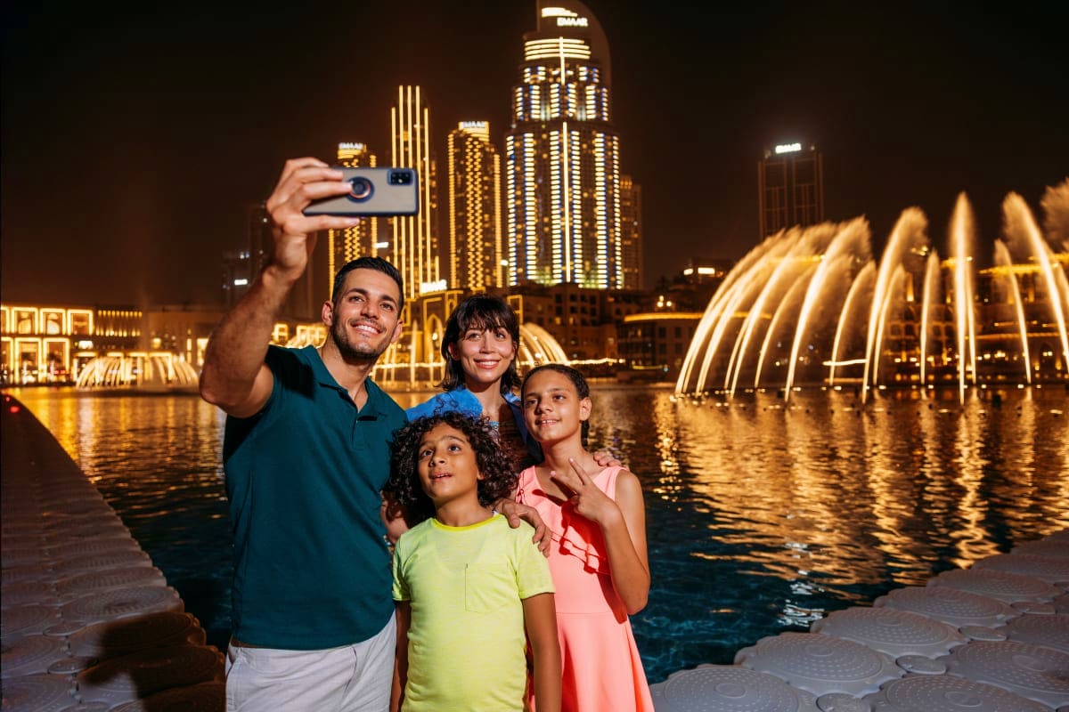 Dubai Fountain Show and Lake Ride Family taking selfie at Dubai Fountain with illuminated skyscrapers and fountain jets reflected in water at night