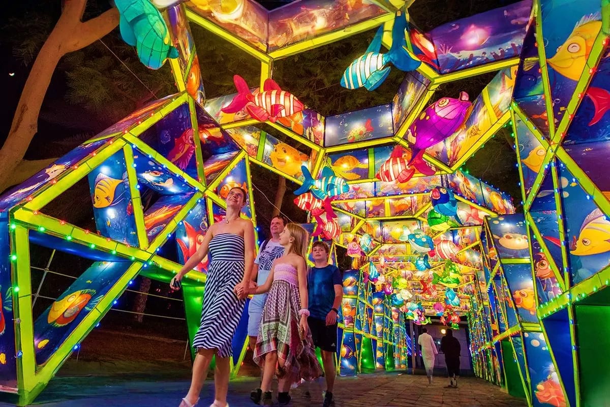 Family walking through illuminated tunnel with neon yellow framework and glowing colorful fish artwork panels at night