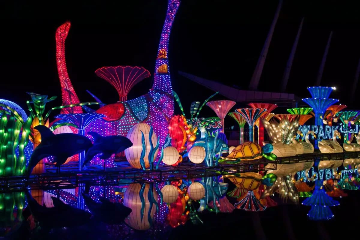 Glow Garden Illuminated lantern sculptures of ocean creatures and plants reflected in water at night, featuring colorful LED-lit whale, j
