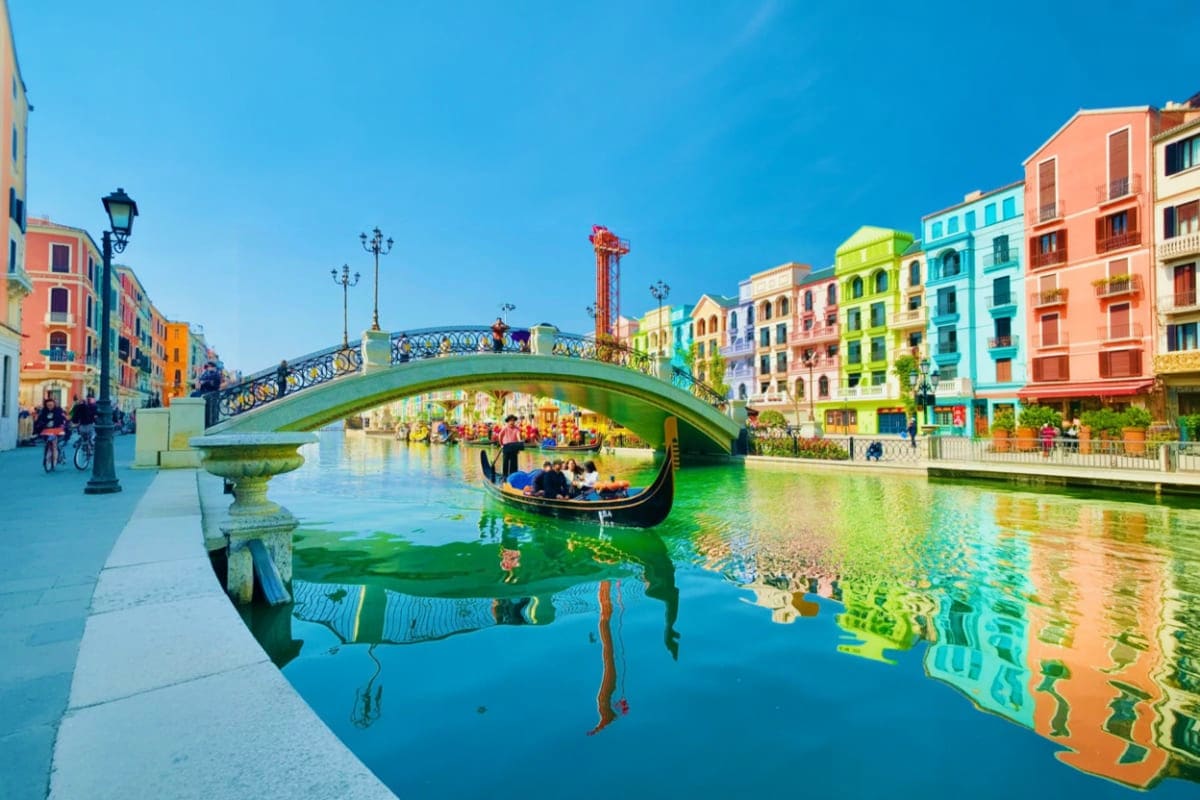 Grand World with Gondola Ride Gondola gliding under arched bridge in Venice-inspired canal with colorful buildings reflected in turquoise water