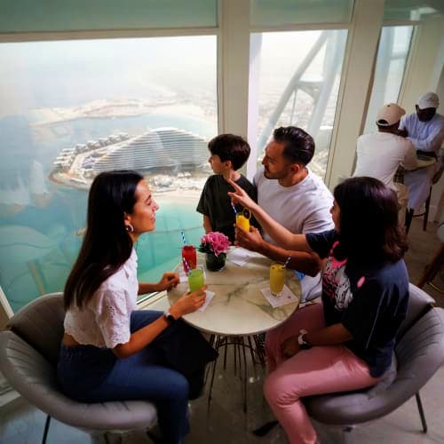 Group of tourists enjoying drinks at a high-altitude interior lounge of Burj Al Arab with panoramic aerial view of Dubai coas