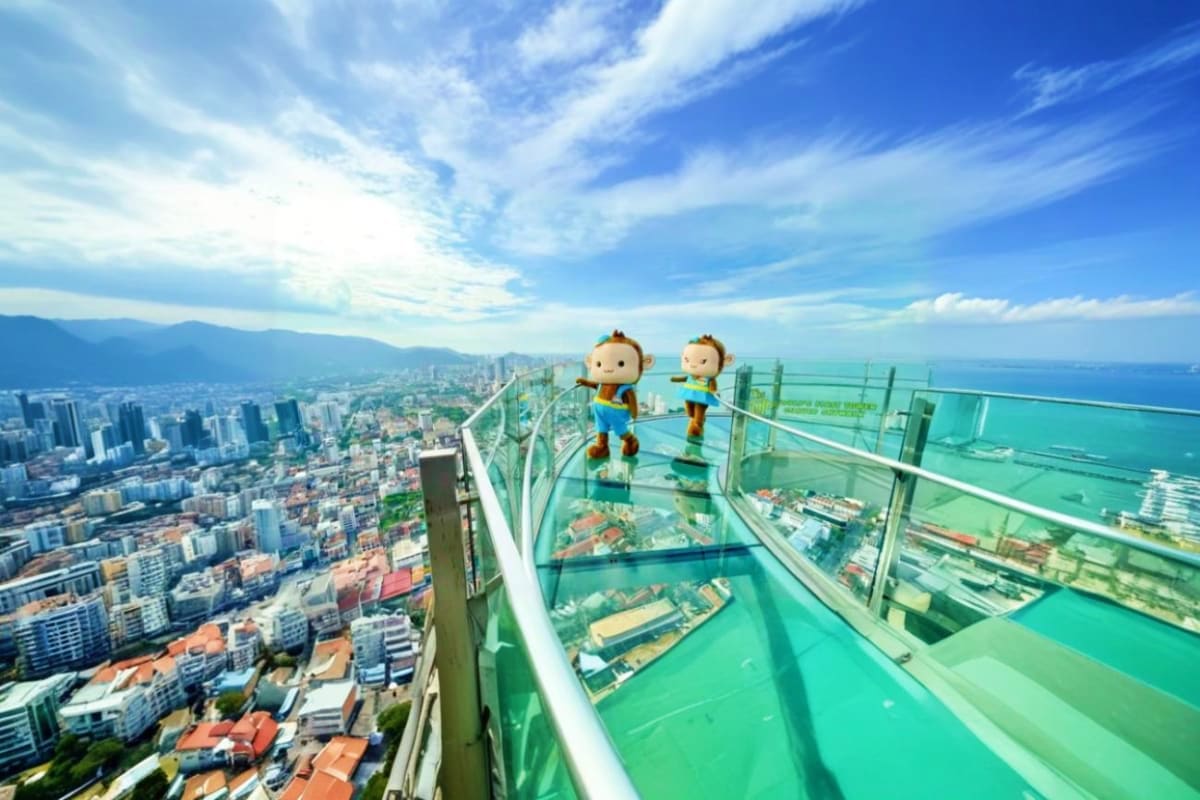 Half Day Penang City Tour Two cartoon characters standing on transparent glass skywalk high above Penang cityscape with mountains and sea in background