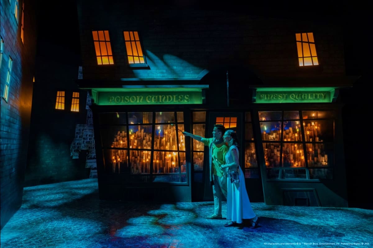 Harry Potter Visions of Magic Two figures outside Ollivander's Wand Shop at night, illuminated by blue lighting and glowing candles visible through storefr