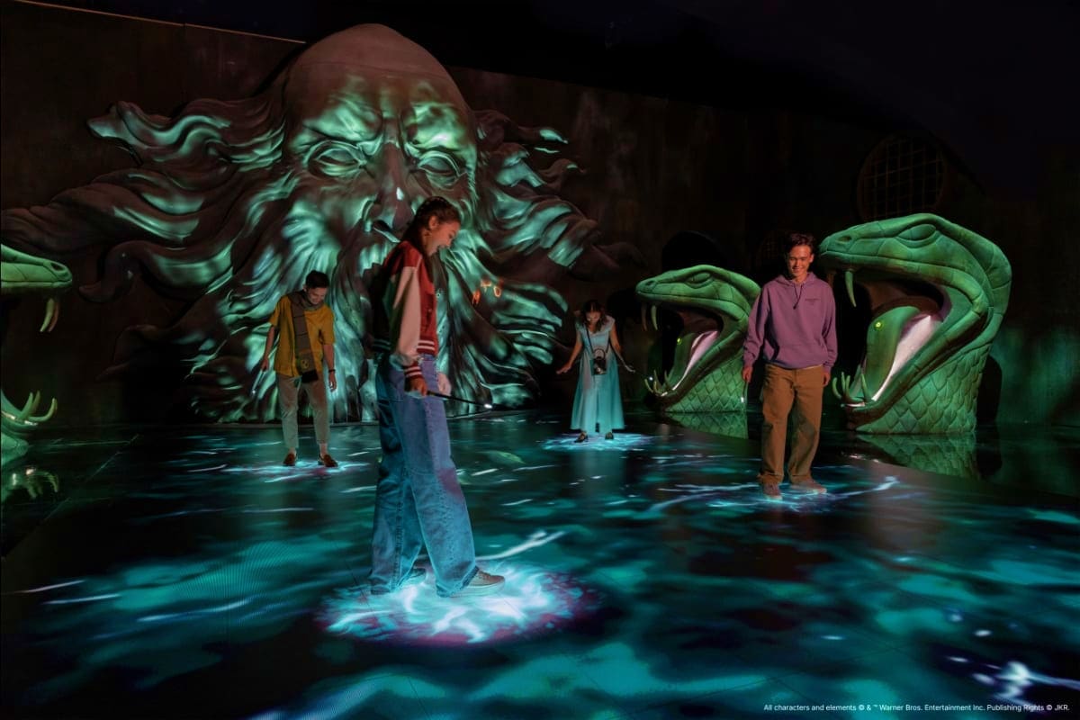Group of visitors standing in immersive magical chamber with glowing water floor and giant green sculptured creature heads lo