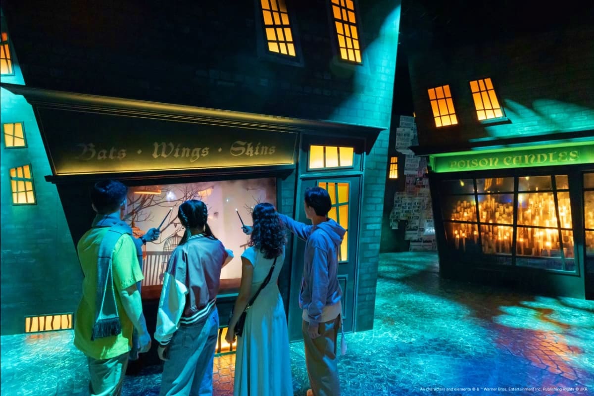 Group of four visitors viewing a display window labeled "Bats Wings Skins" in a themed street with glowing yellow windows and