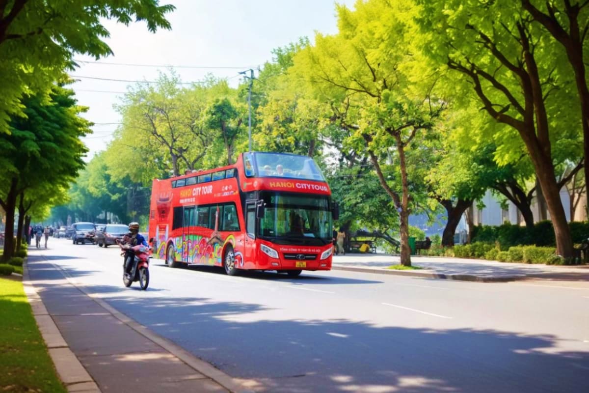 Red double-decker Hanoi City Tour bus driving down tree-lined street with motorcyclist and pedestrians nearby