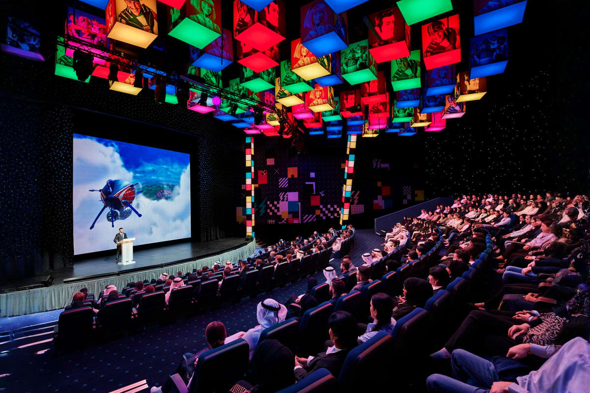 IMG Worlds of Adventure Audience seated in a modern theater with colorful illuminated ceiling panels watching a presentation on a large screen displa