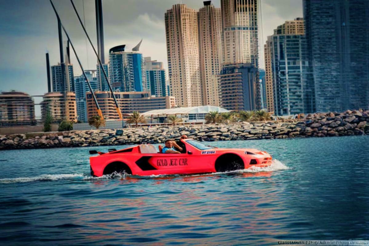 Red amphibious jet car driving in turquoise water with Dubai Marina high-rise buildings and rocky breakwater in background